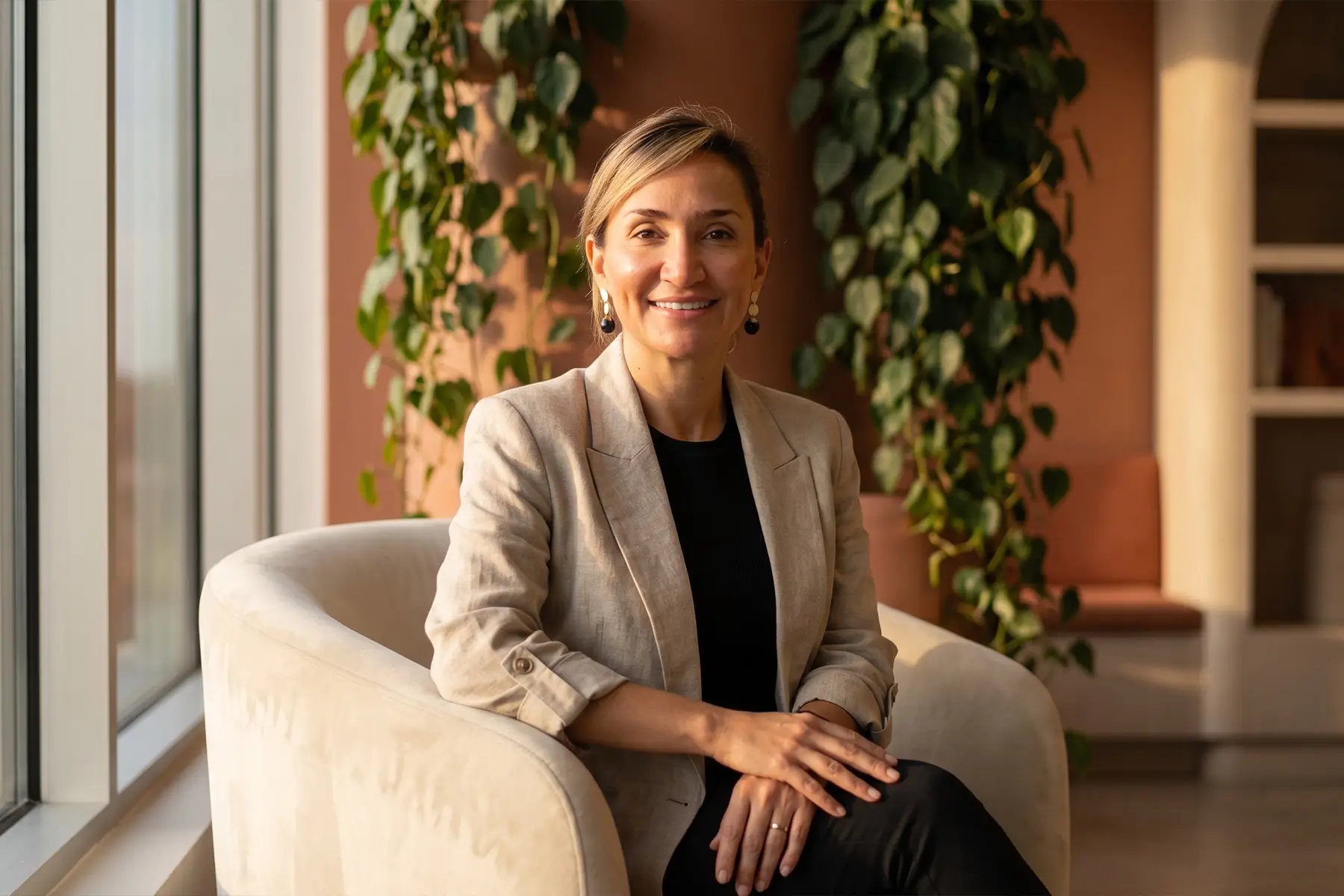 Close-up professional portrait of Business Unit Director Bruna Esposito smiling warmly in a cream armchair, with large trailing green plants and terracotta walls in the background.
