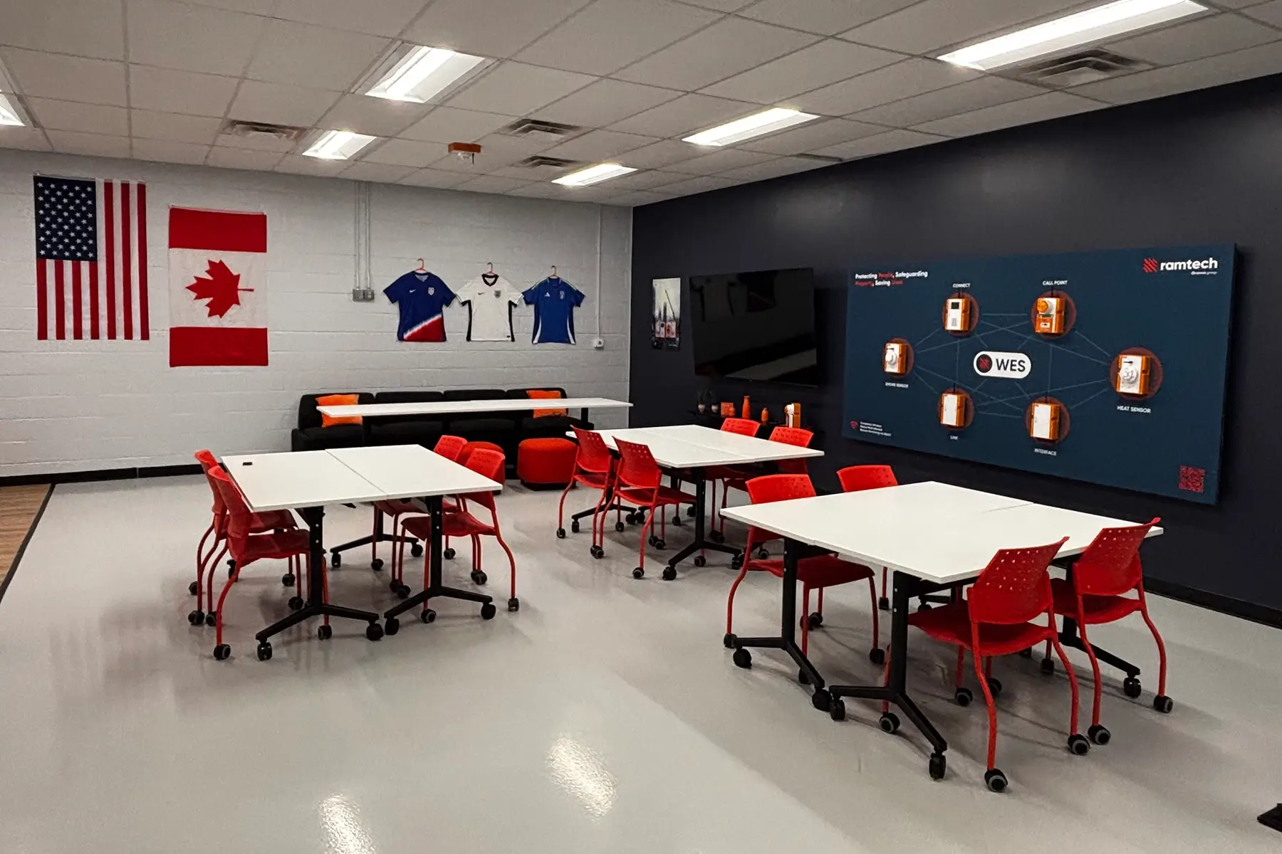 Training room at the Ramtech Charlotte office with tables, red chairs, flags, jerseys, and a screen displaying the WES system network.