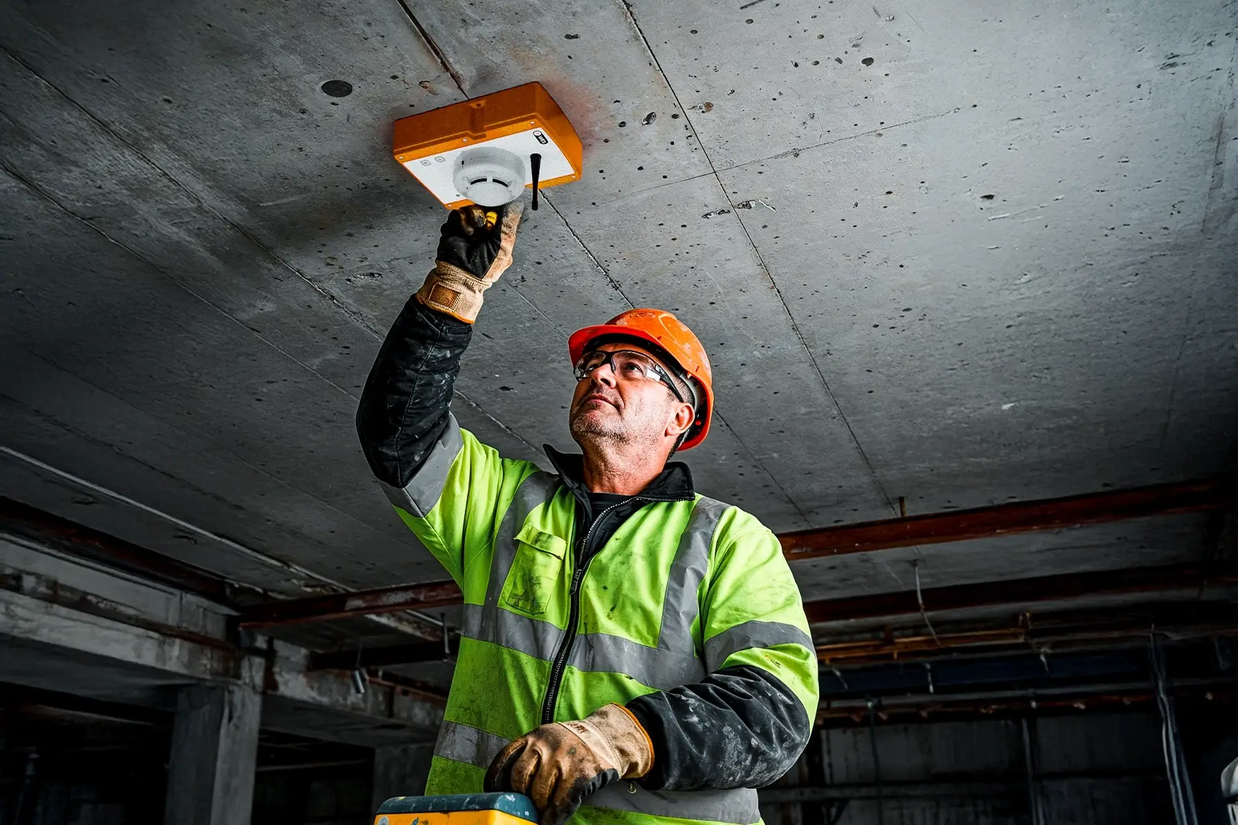 A worker wearing a hard hat and high-vis jacket installs a detector that is part of the WES wireless life safety system on a raw concrete ceiling.