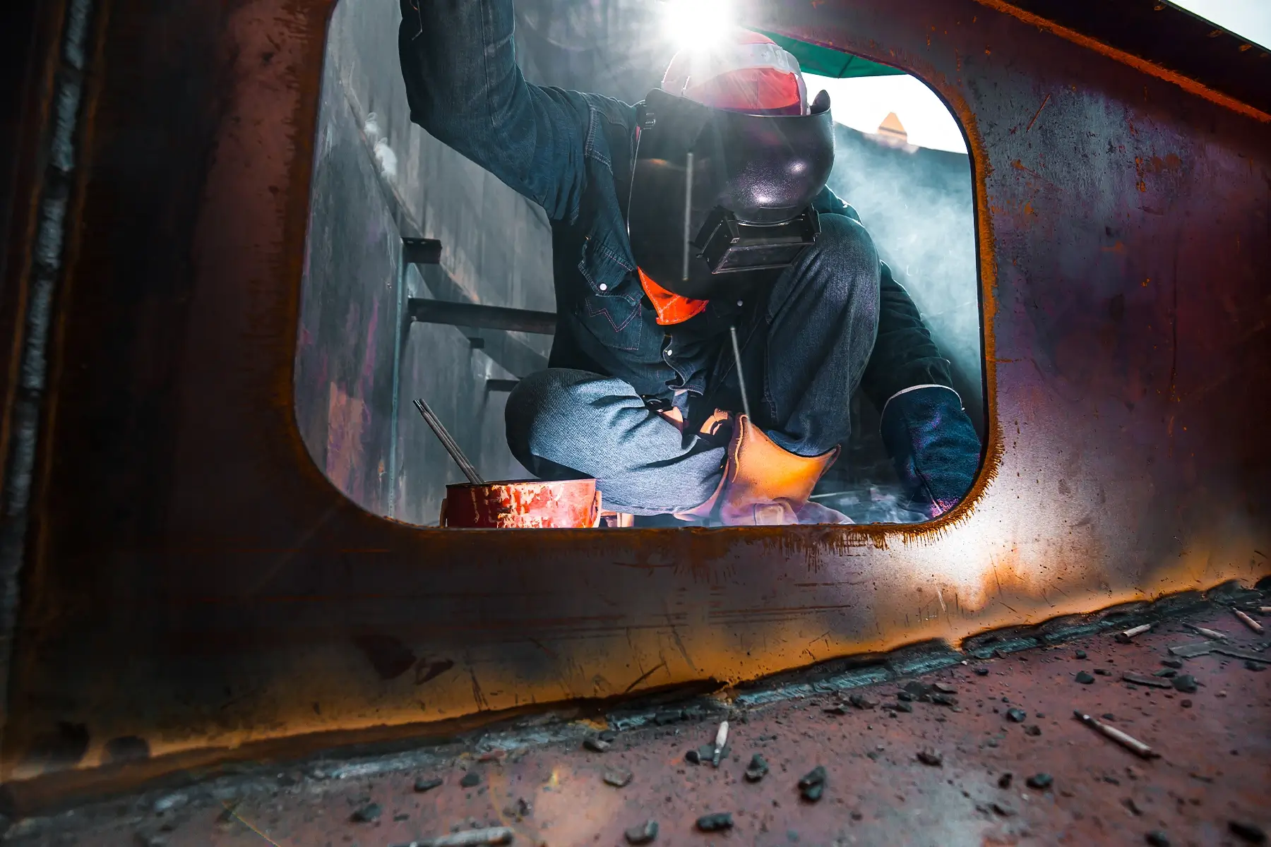 A welder working in a dark, confined space with an orange glow, demonstrating high-risk hot-works activity requiring a WES wireless life safety system.
